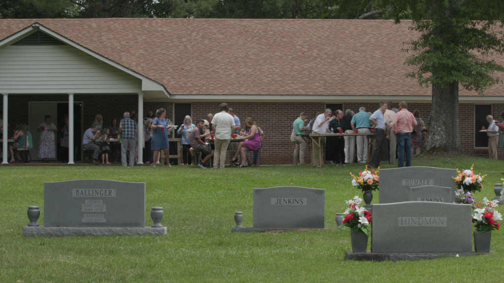 Photograph of people gathered around tables eating outside a church building; gravestones with the names Ballinger and Jenkins in foreground.