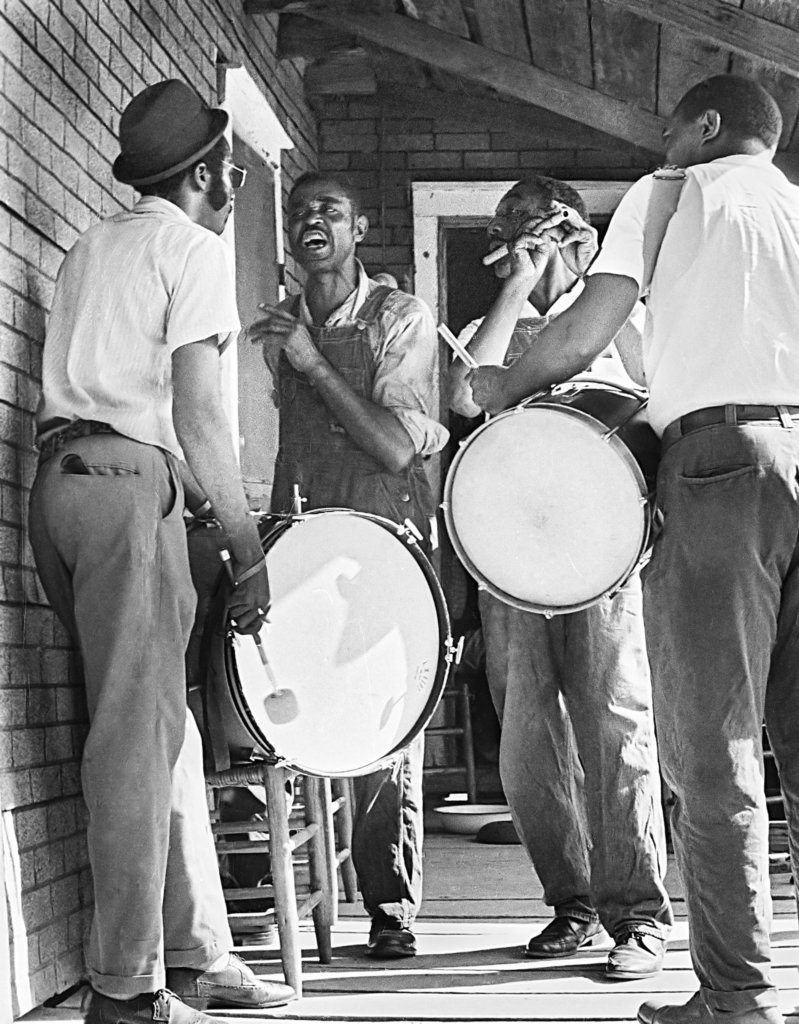 Four musicians on a porch, one singing, two playing snare drums, and one playing a flute.