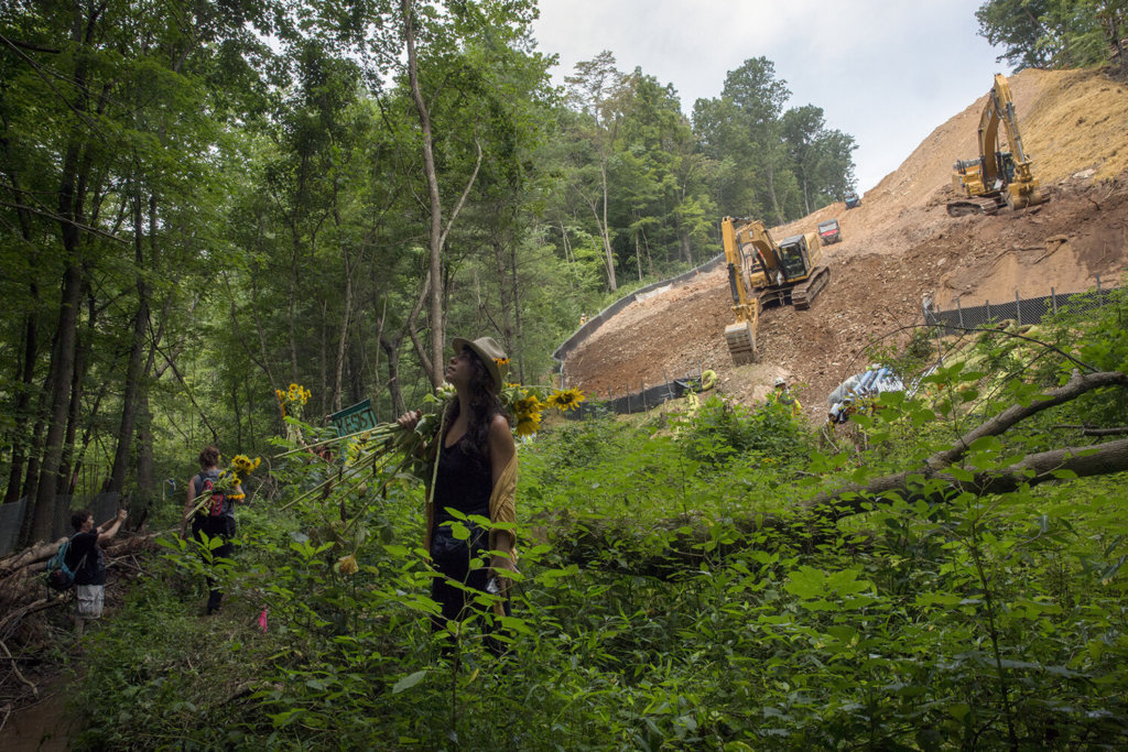 Laney Sullivan and other protestors stand in a forested area with bulldozers and developed land behind them.