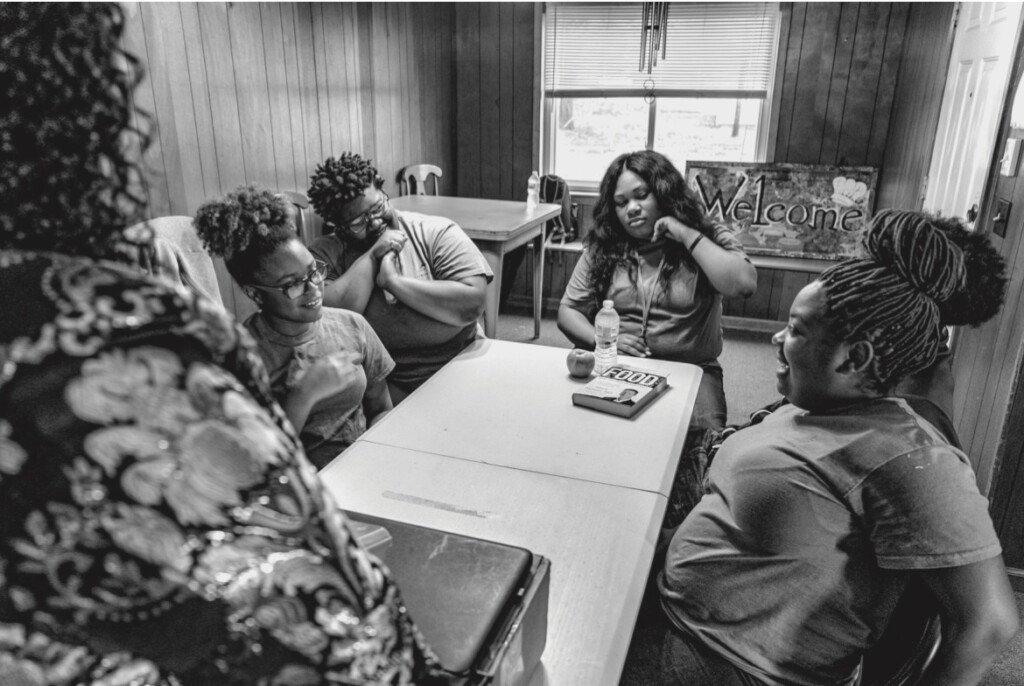 Looking over the shoulder of an adult woman whose face is not in frame, four young African American teenagers sit around a small table, smiling and talking; an apple, bottle of water, and a book with the word "Food" sits closet to the young woman by the window.