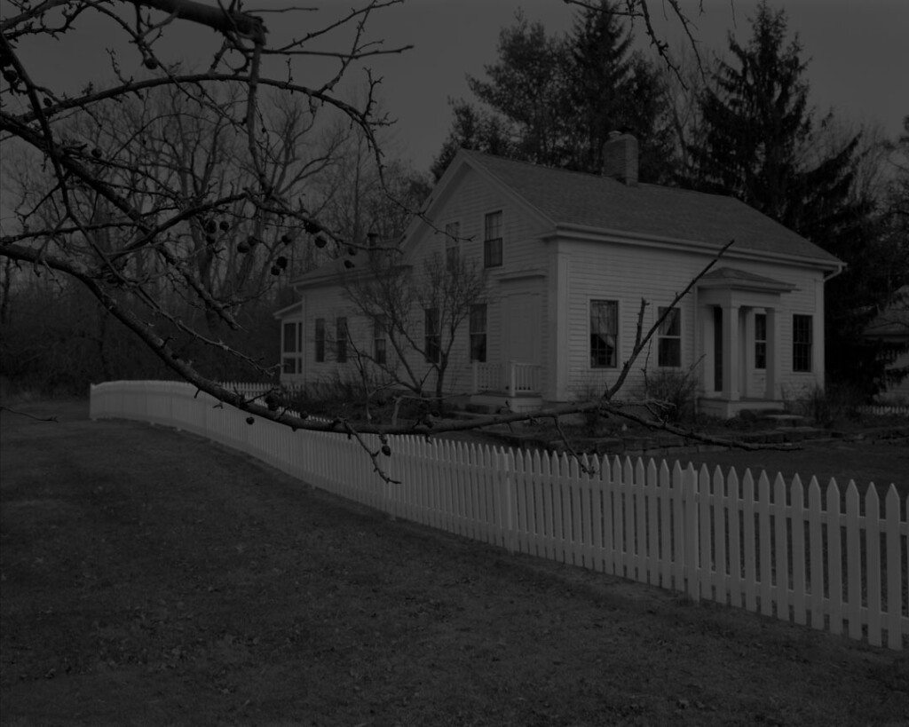 A large house stands behind a long white picket fence. Barren branches from the surrounding trees covers the façade in the foreground.