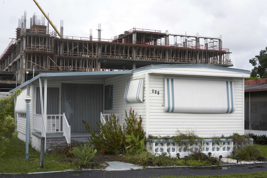 Foreground a well-tended double-wide of white planks and blue trim while in the background a monstrous banality of rebar rises as if to devour the trailer in one rapacious gulp.
