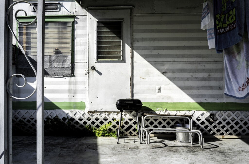 The horizontal lines of jalousie windows on a mobile home stand perpendicular to the vertical lines of a metal post and door frame. Both then seem to merge with criss-cross lattice, as a clearly delineated triangle of shade falls across the trailer.