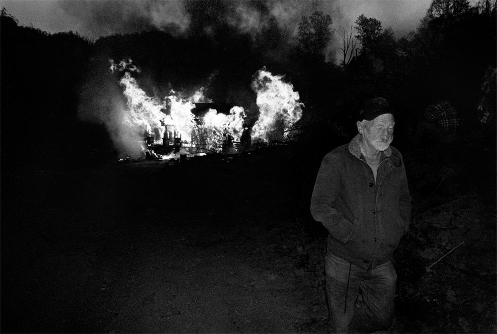 Older man walking away from burning house at night.