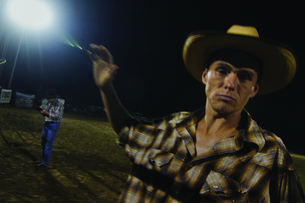 Young man at rodeo with cowboy hat on and one arm raised toward camera.